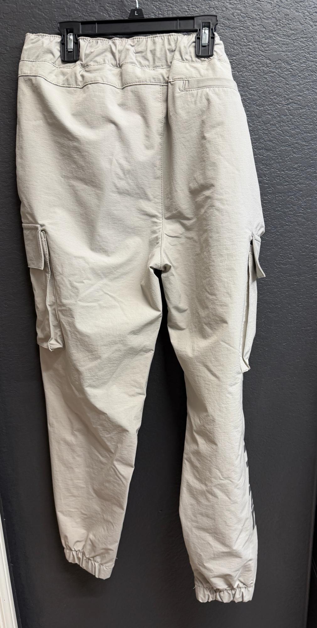 Beige cargo pants with an elastic waistband and side pockets, hanging on a black hanger with the letter L, over a dark gray wall.