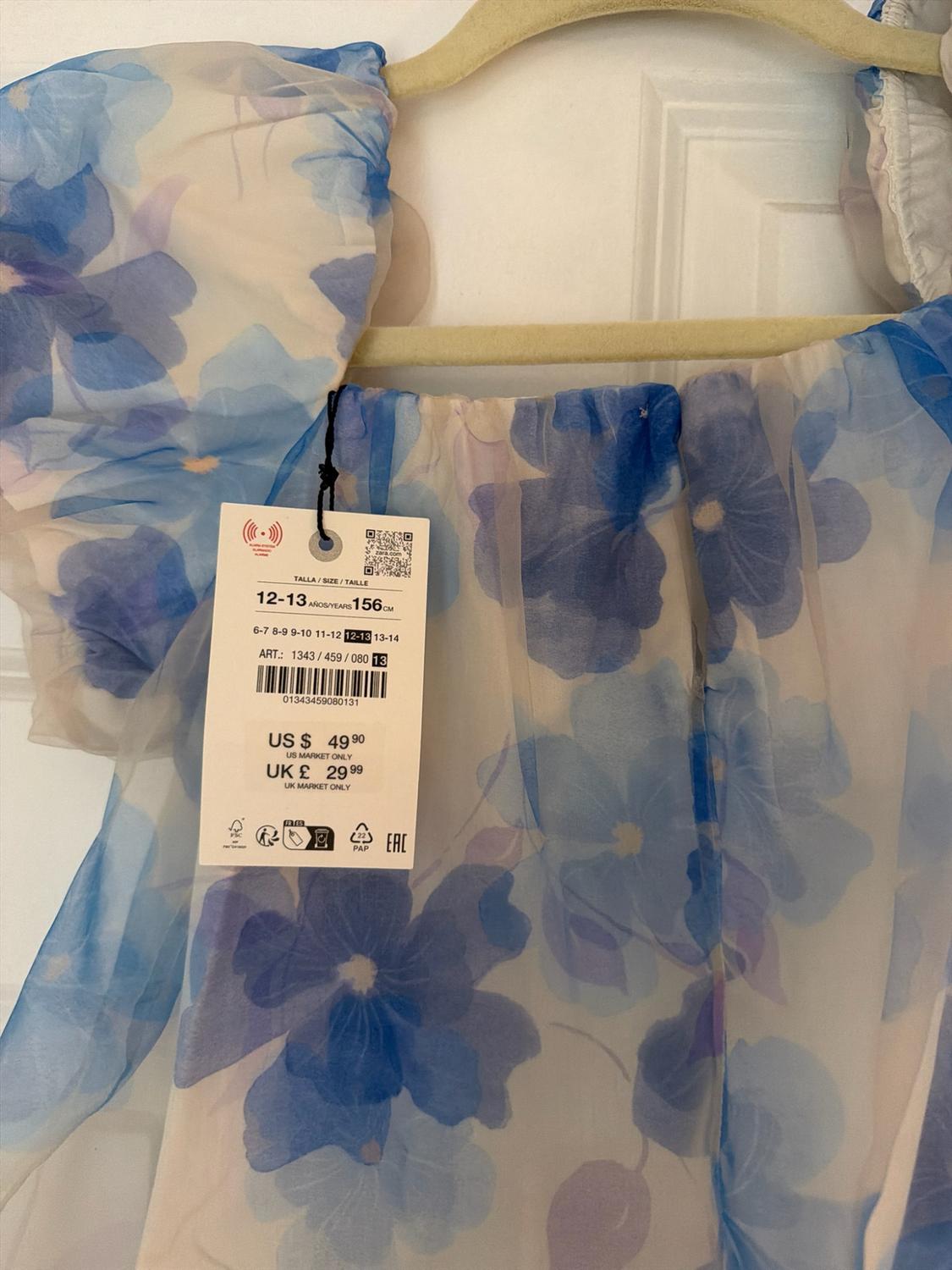 White dress with a blue floral print and puff sleeves on a hanger. Label: 12–13 YEARS 156 CM US $49.90 UK £29.99.