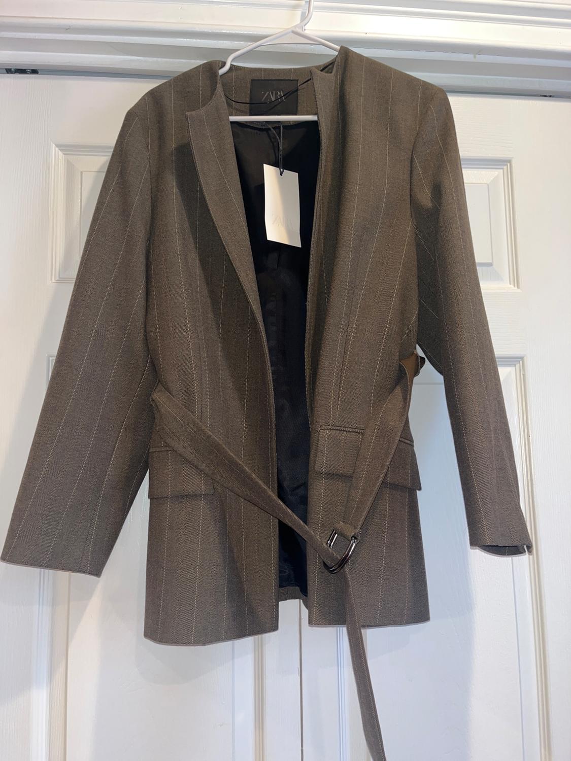 Brown blazer with fine stripes hanging on a white hanger. It has a belt and a visible label inside. White door background.