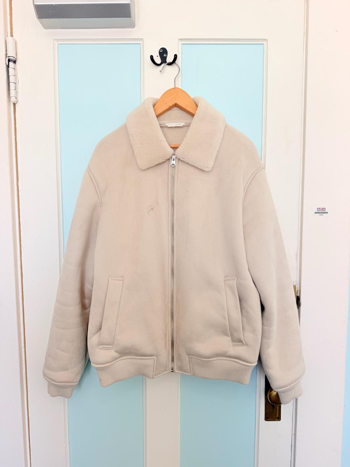 Casual beige jacket with a zip, pockets, and a fleece collar, hanging on a wooden hanger on a white door with blue panels.