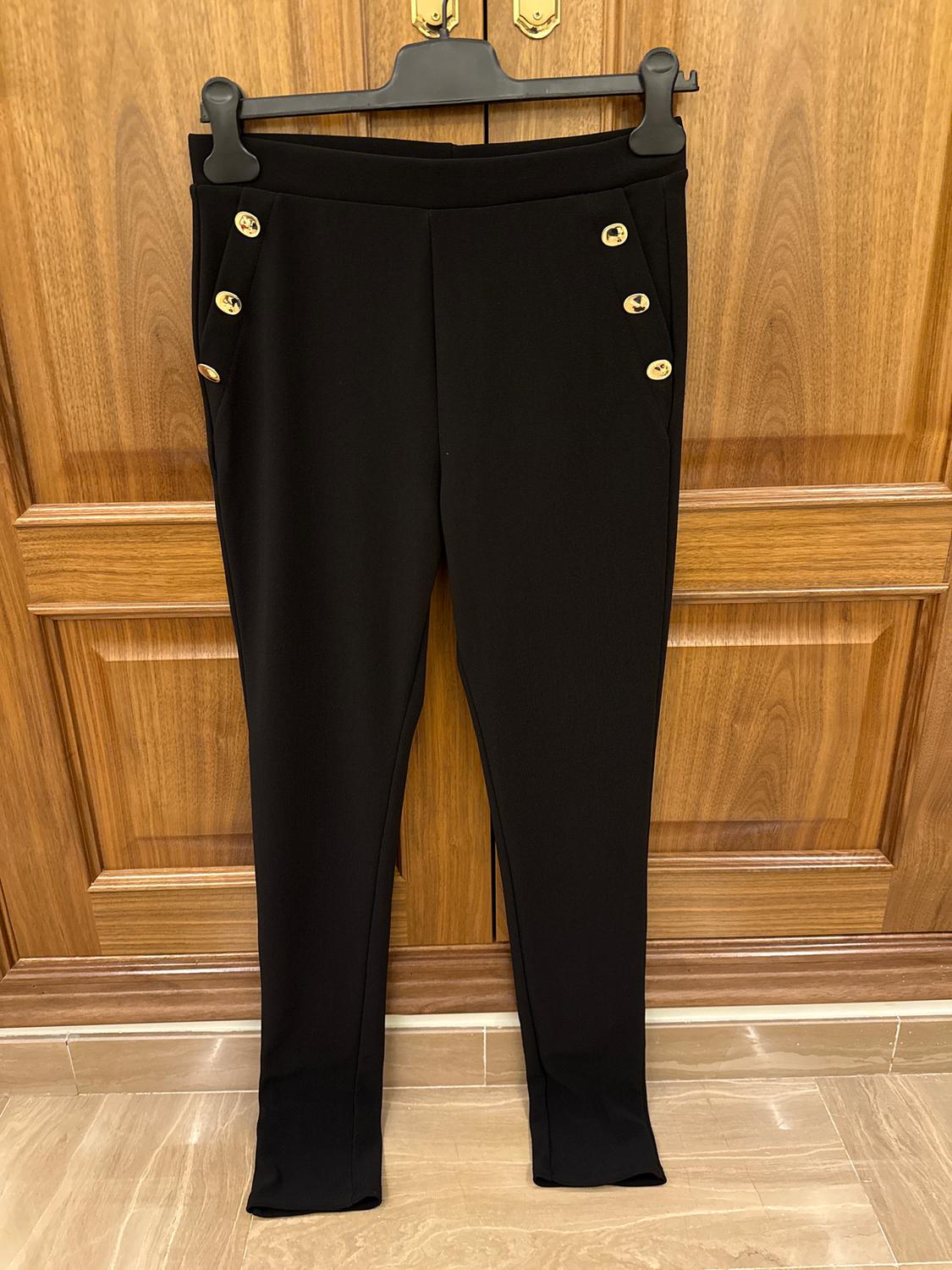 Black trousers hanging on a hanger, with gold buttons on the waistband, against a brown wooden background.