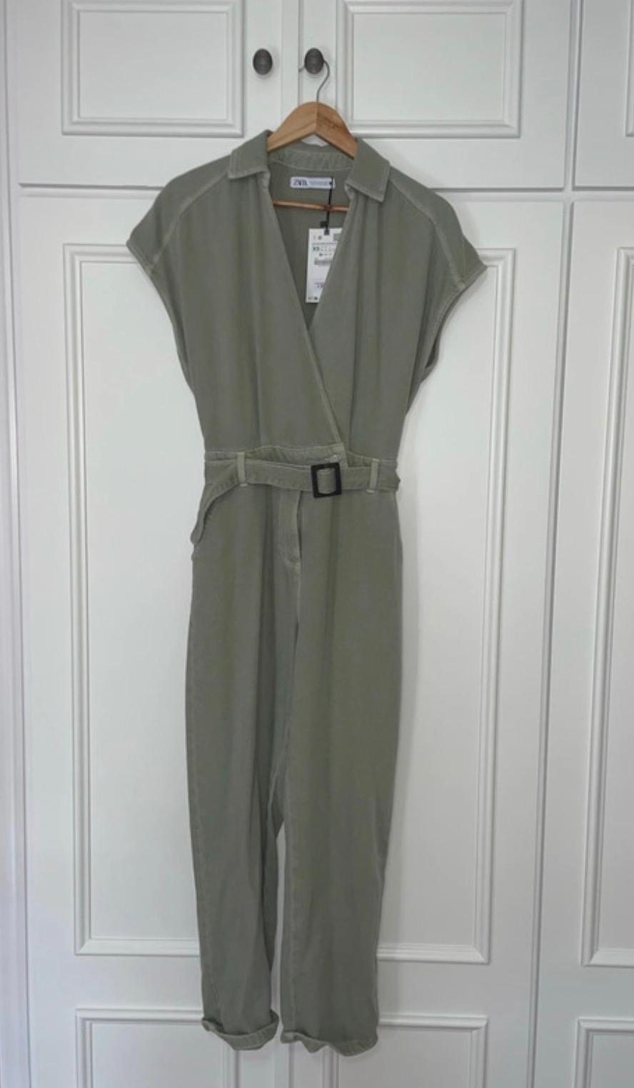 Olive green jumpsuit with black belt hanging on a wooden hanger in front of a white paneled wardrobe.