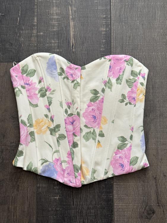 Floral corset with a print of pink, lilac, and yellow roses on a cream background, placed on a dark wood floor.