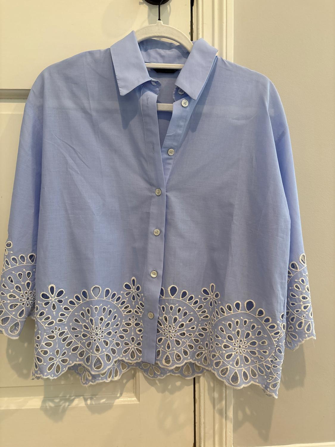 Light blue button-down shirt with white openwork embroidery on the hem and cuffs, hanging on a hanger over a white door.