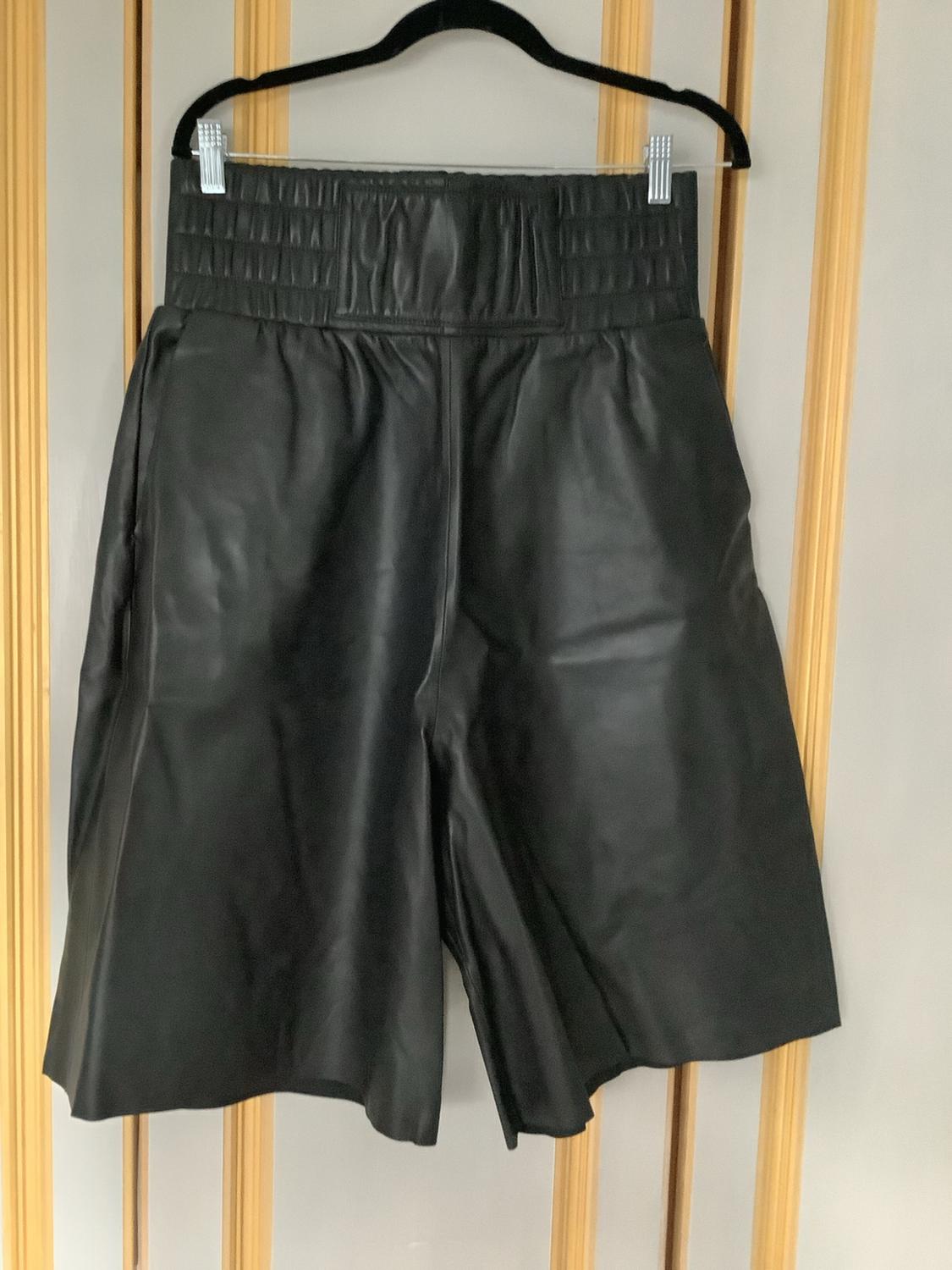 Black leather Bermuda shorts with a wide elasticated waistband, hanging on a black hanger in front of a wall with light wood slats.
