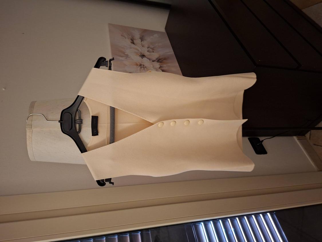 Sleeveless beige waistcoat hanging on a mannequin, with front buttons, against a white wall and dark furniture background.