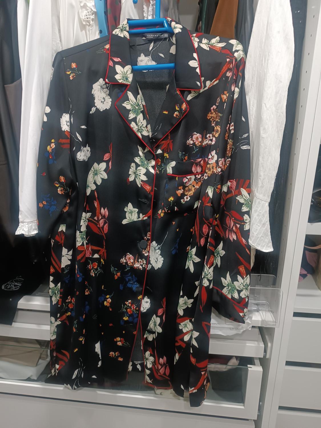 Black satin shirt with a multicoloured floral print and red piping, hanging in a wardrobe next to other garments.