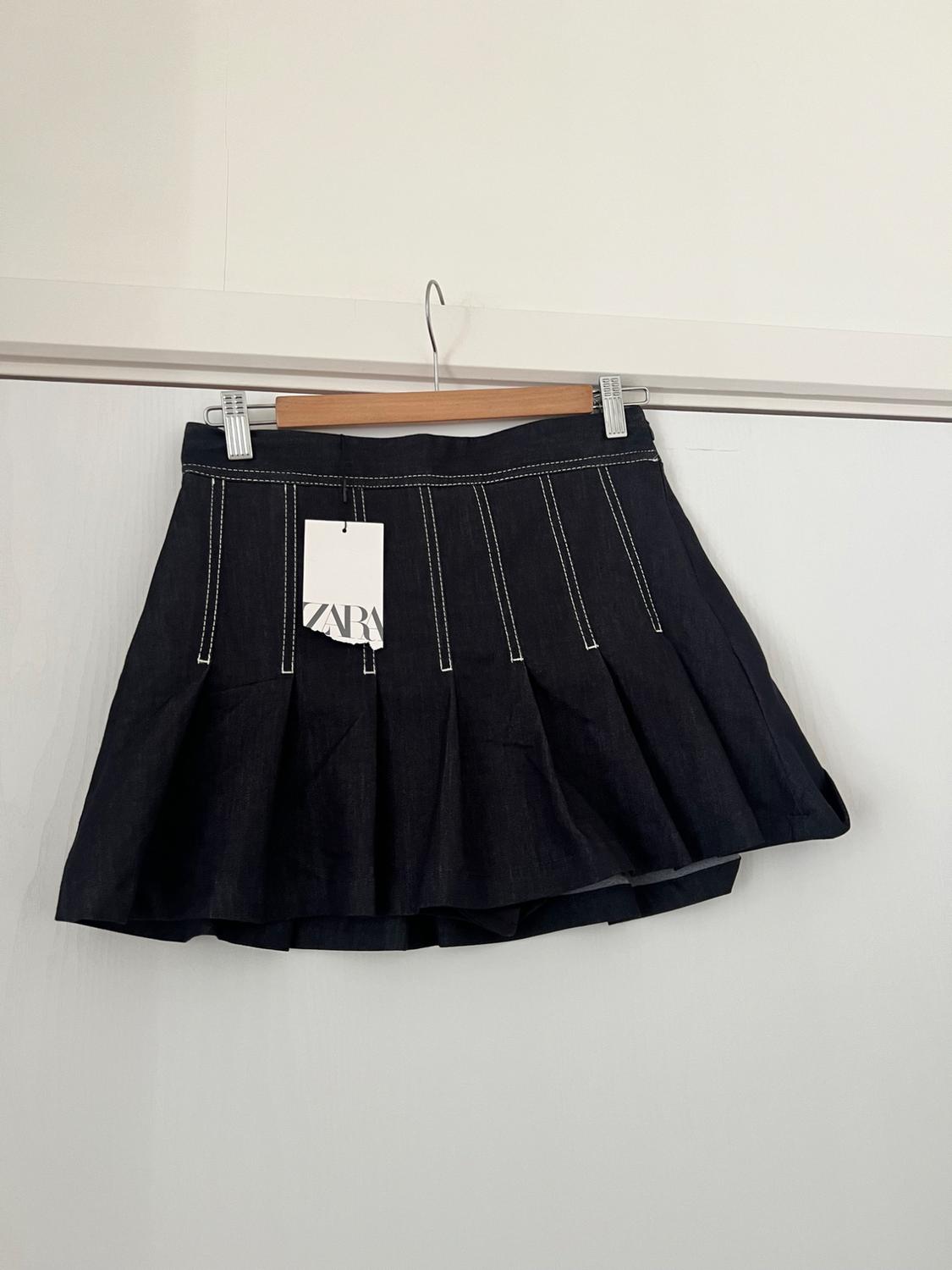 Dark denim pleated mini skirt with white stitching, hanging on a wooden hanger against a white background. Label with the text ZARA.