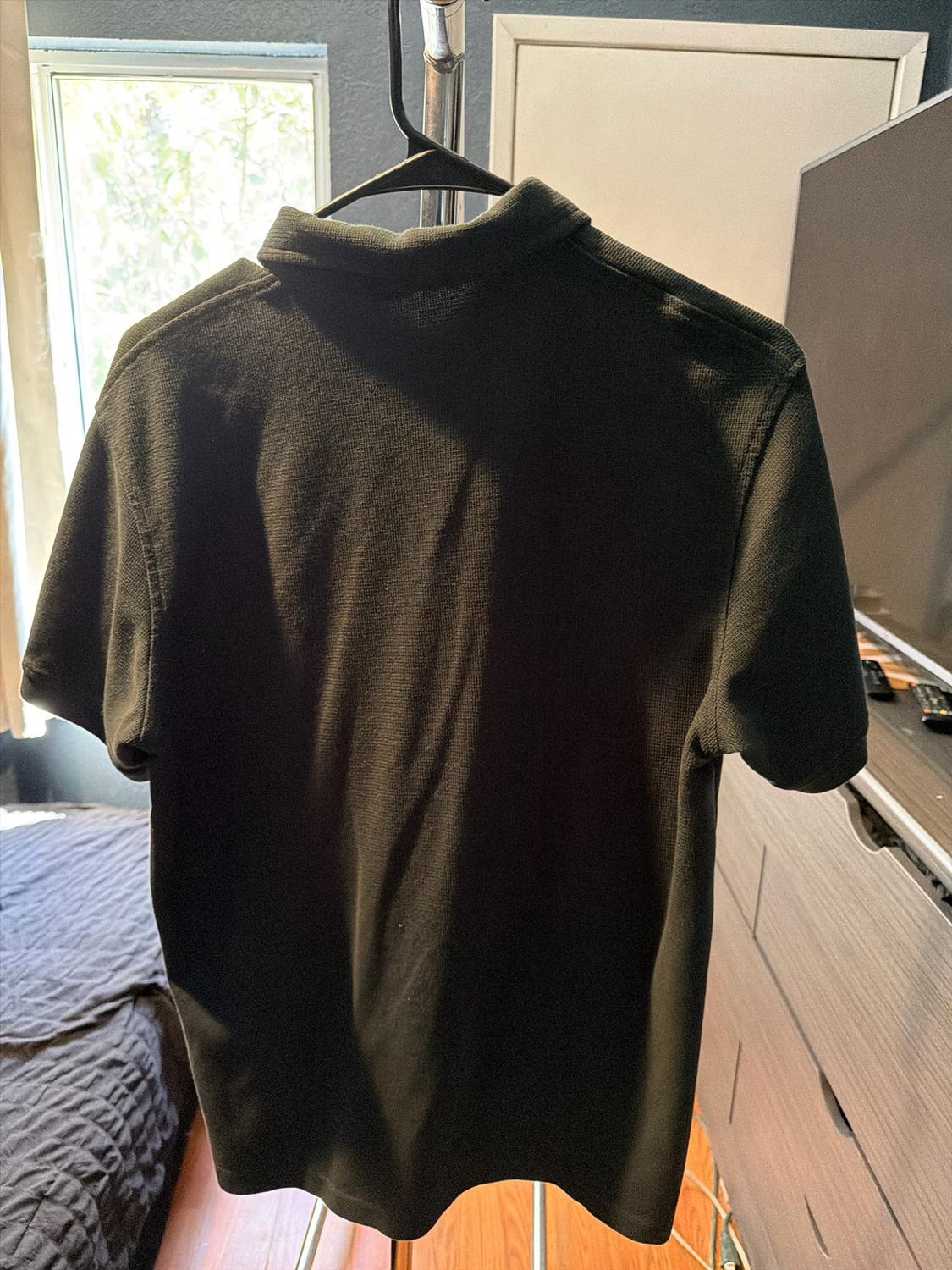 Back of a dark short-sleeved polo shirt hanging on a hanger in a bedroom with natural light, a bed, and wooden furniture.