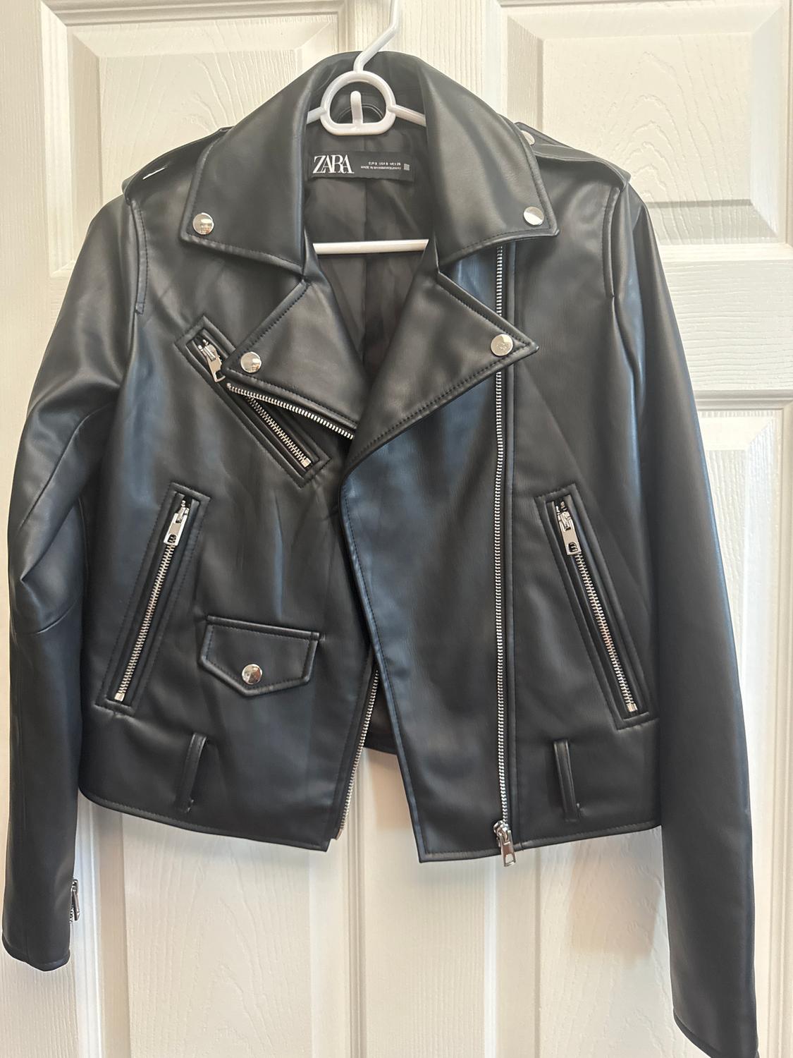 Black faux leather jacket with zips and silver buttons, hanging on a white hanger in front of a white door.