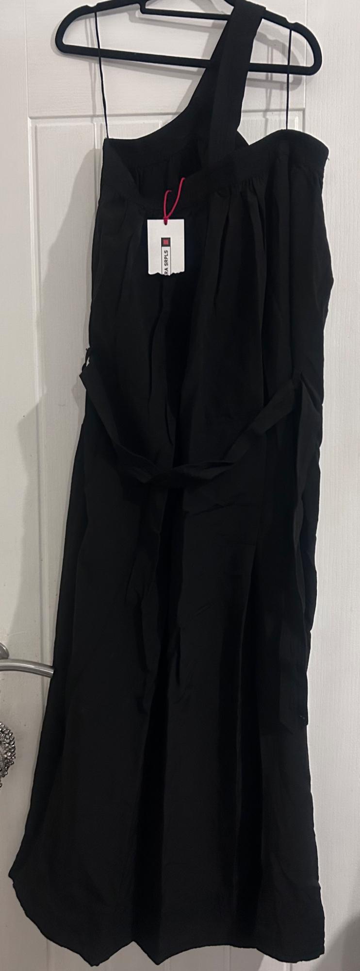 Black one-shoulder dress hanging on a black coat hanger. Visible label with text "SIRPLUS". White door background.