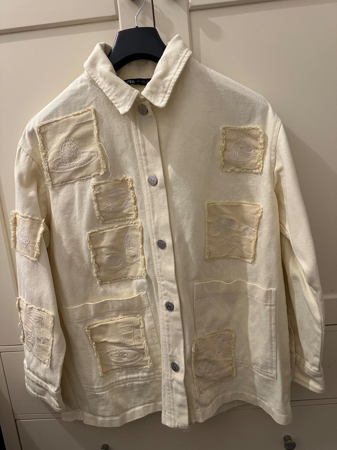 Beige jacket with raised embroidered patches, hanging on a black hanger. White wardrobe background.