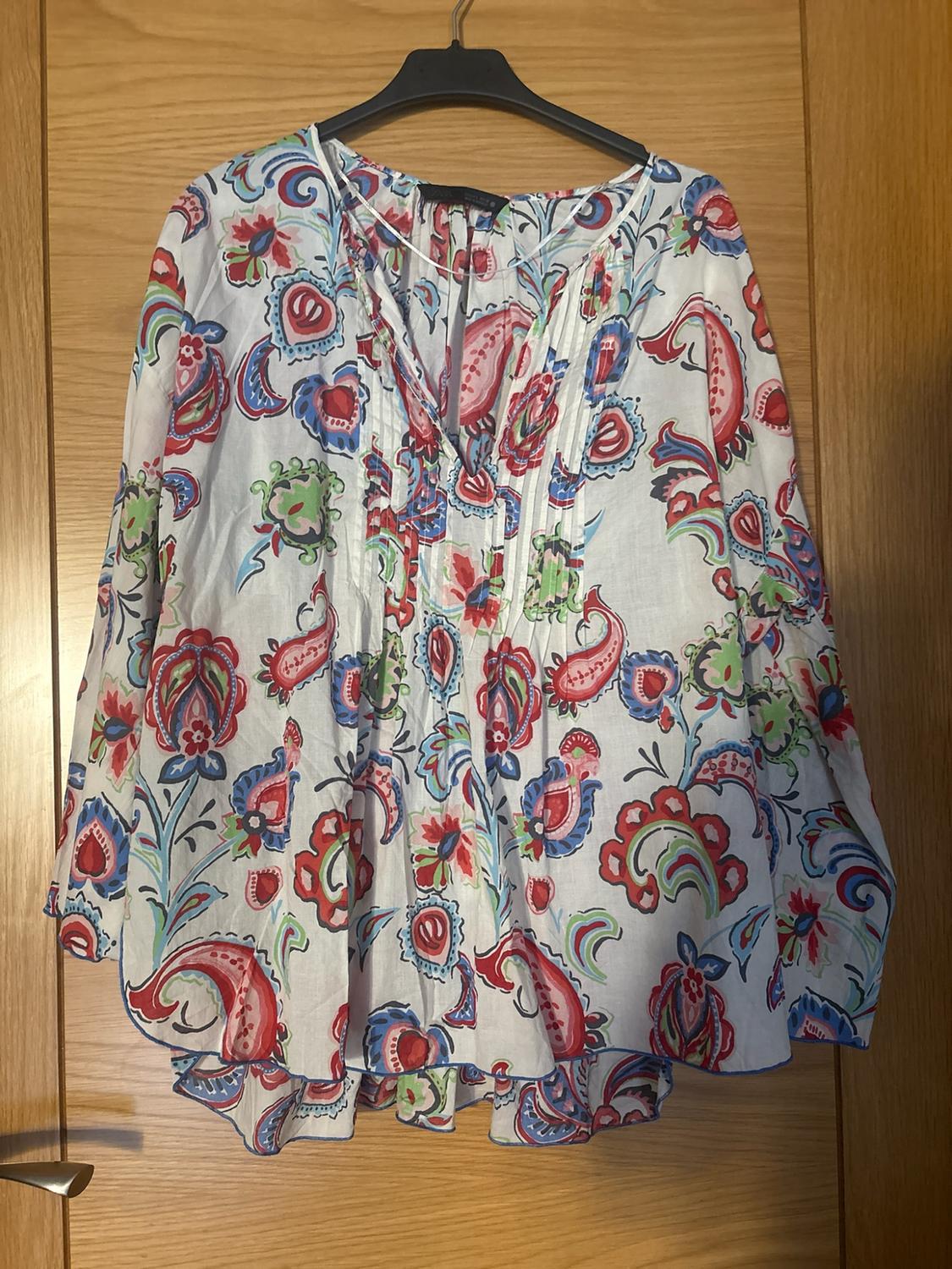 White blouse with a multicoloured red, blue and green print, V-neck and front pleats, hanging on a wooden door.