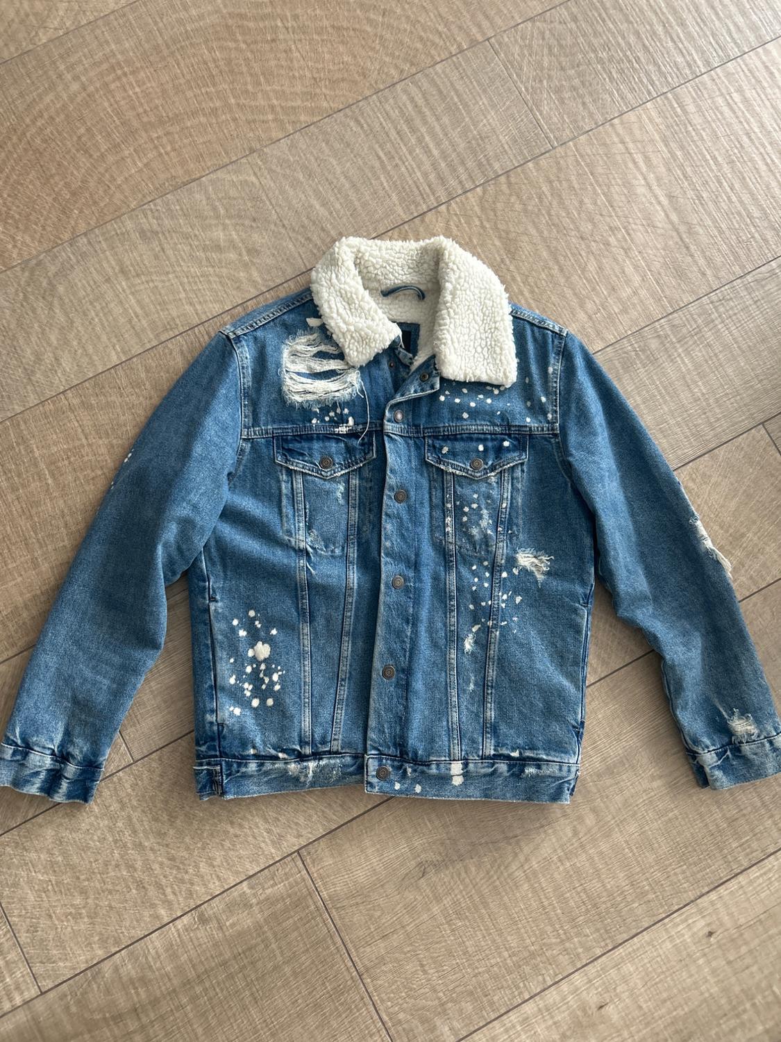 Blue denim jacket with a wool collar, distressed details, and white splatters, on a light wooden floor.