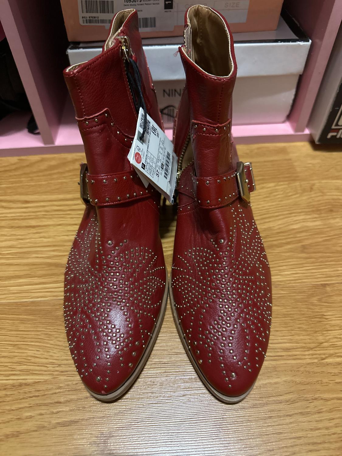 Red leather ankle boots with gold studs and buckles, on a wooden floor. Price tag visible on one of the boots.