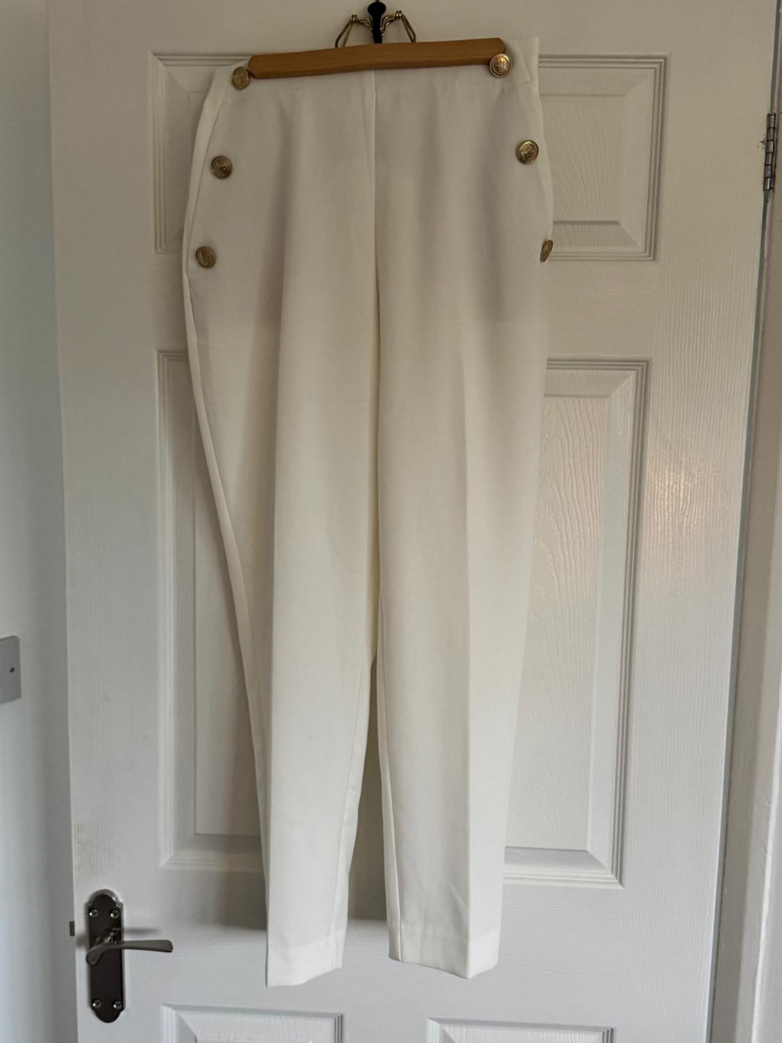 White, lightweight fabric trousers with decorative gold buttons on the sides, hanging on a hanger in front of a white door.