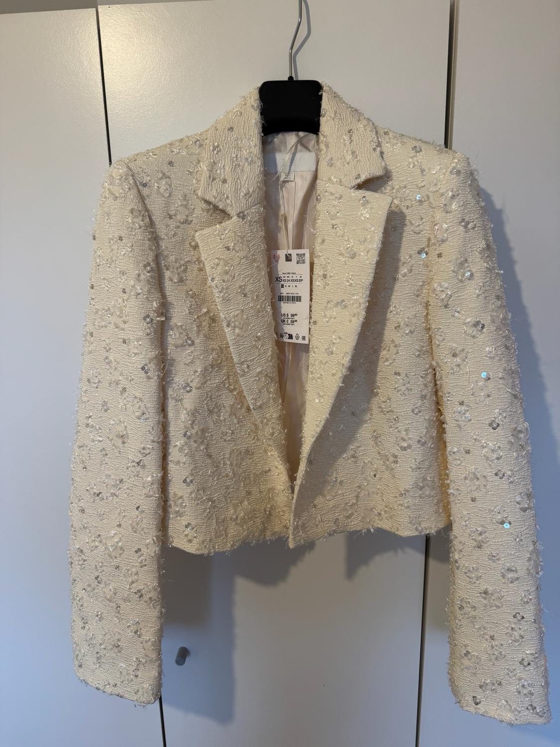 Textured cream sequinned jacket on a black hanger against a white background. Text: XS, US $ 99.90, UK £ 59.99.