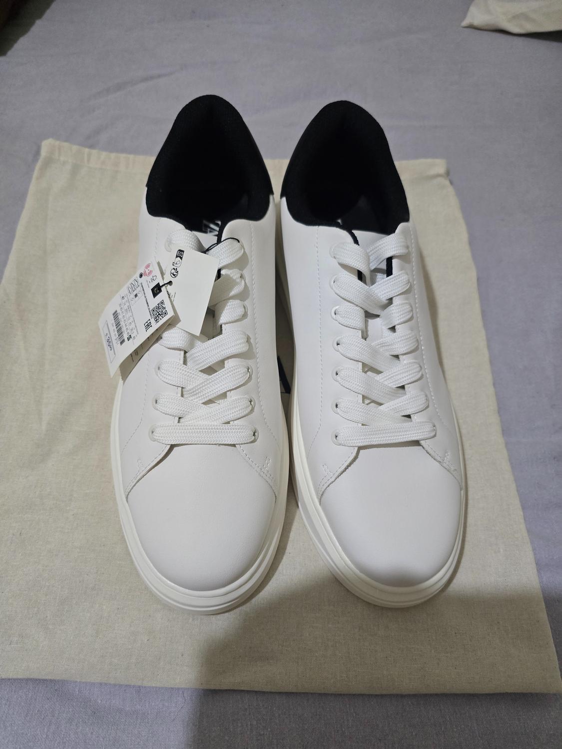 Pair of white trainers with laces and a black interior, resting on a light-coloured fabric bag. The left trainer has a label.