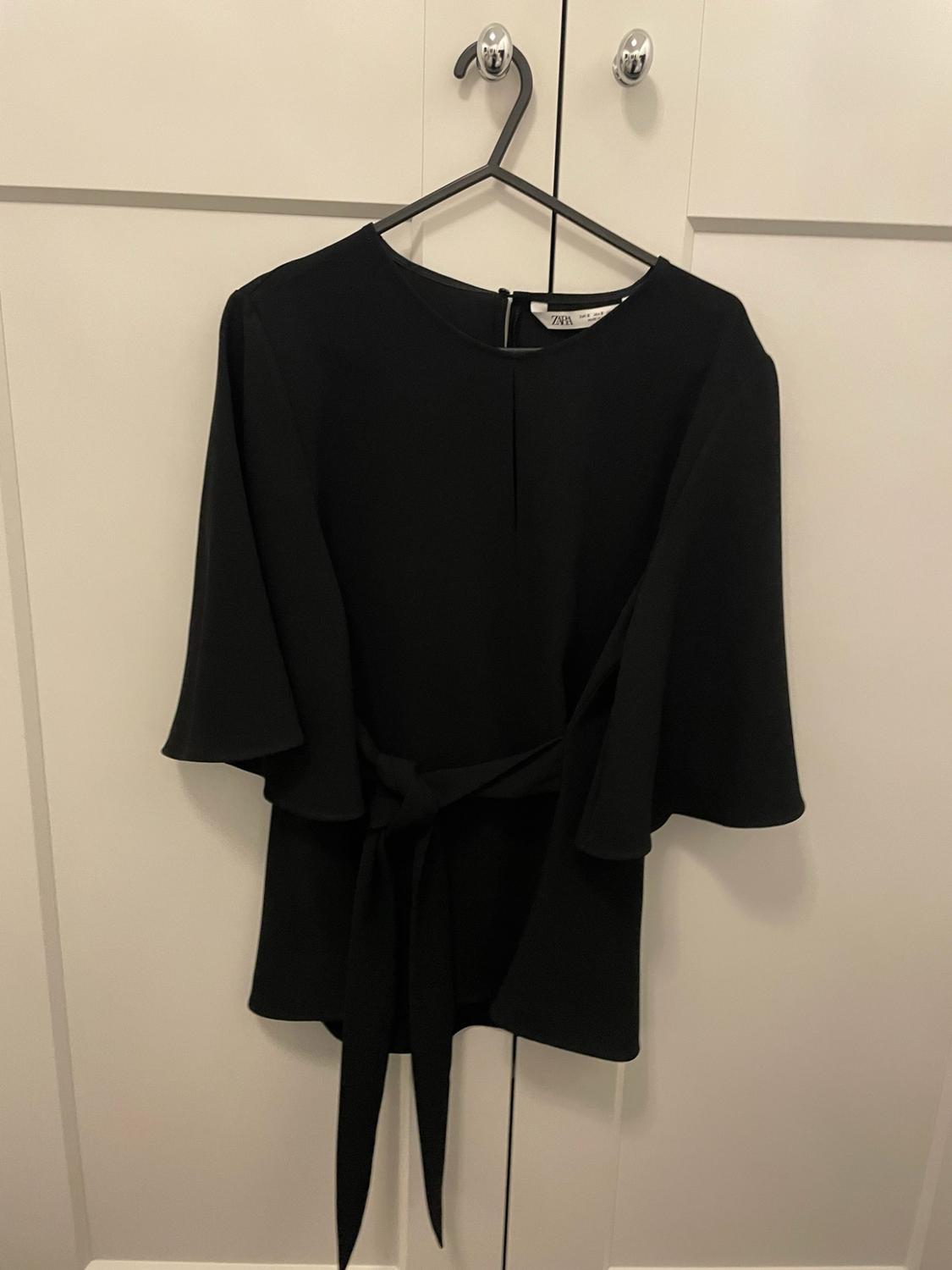 Black blouse with wide sleeves and a tie waist, hanging in a white wardrobe. ZARA text on the label.