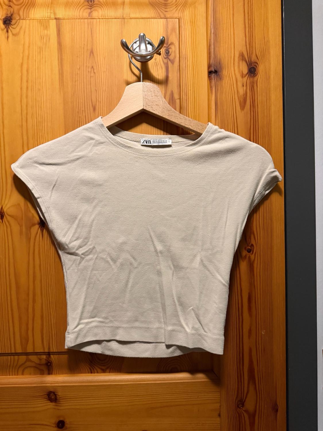 Basic beige cropped short-sleeved T-shirt hanging on a wooden hanger over a door. The label reads ZARA.