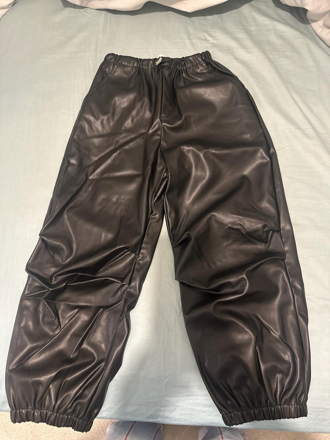 Black shiny jogger-style trousers with an elasticated waist and cuffs, laid out on a light blue sheet.