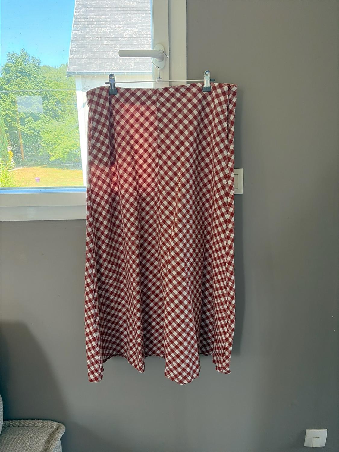 Long red and white checked skirt hanging on a hanger from a window handle. A grey wall and a garden can be seen in the background.