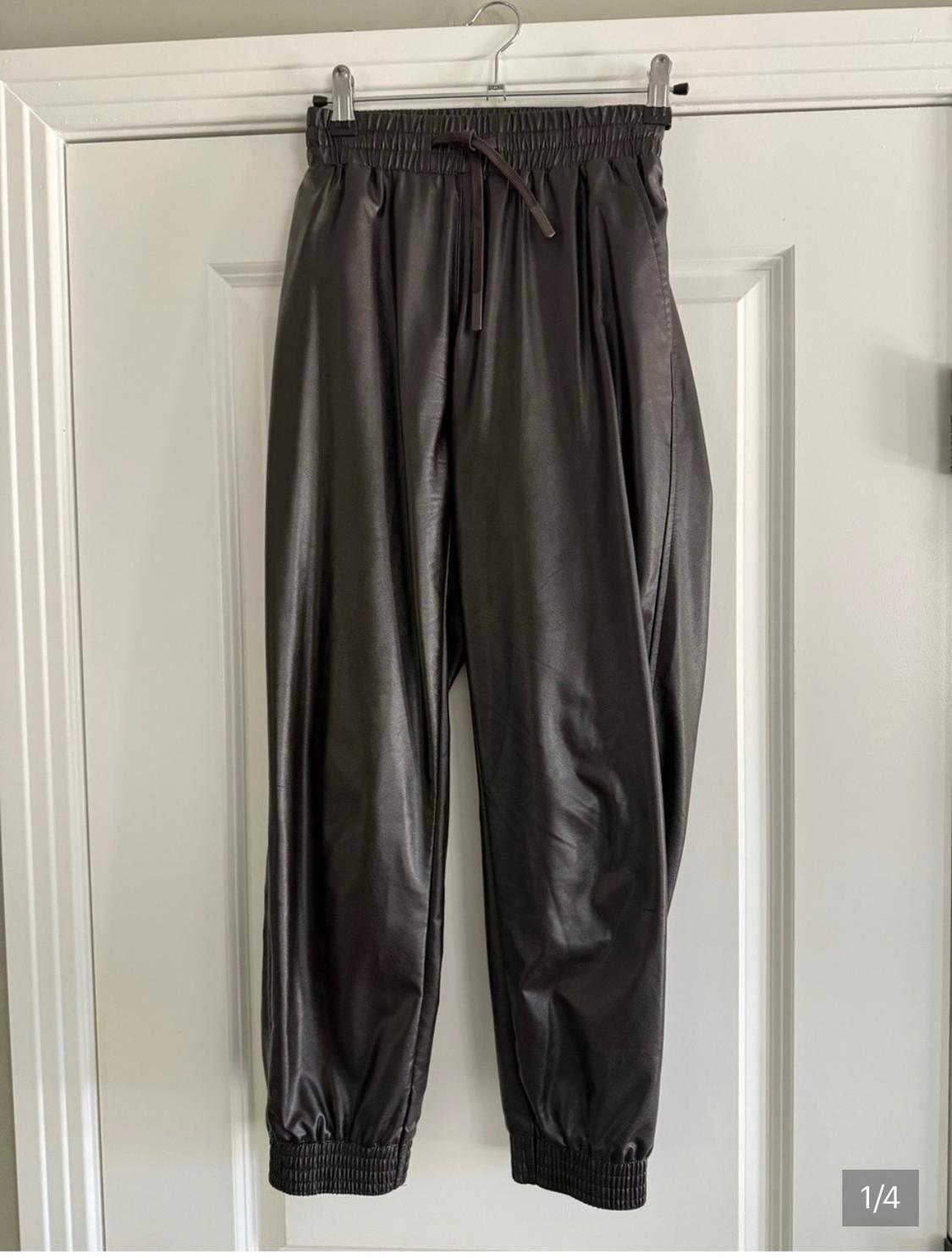 Dark glossy joggers with an elastic waist and drawstring, hanging on a white door. Text: 1/4.