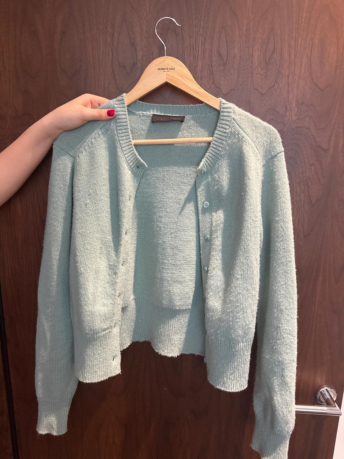 A light blue cardigan is hanging on a wooden hanger with the label "Kenneth Cole". A hand is holding the hanger in front of a wooden door.