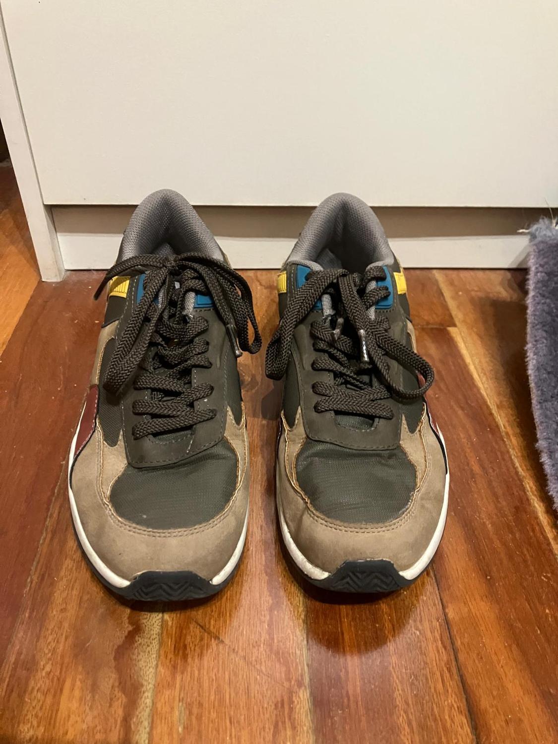 Trainers in brown and green tones with blue and yellow details, on a wooden floor next to a grey rug.