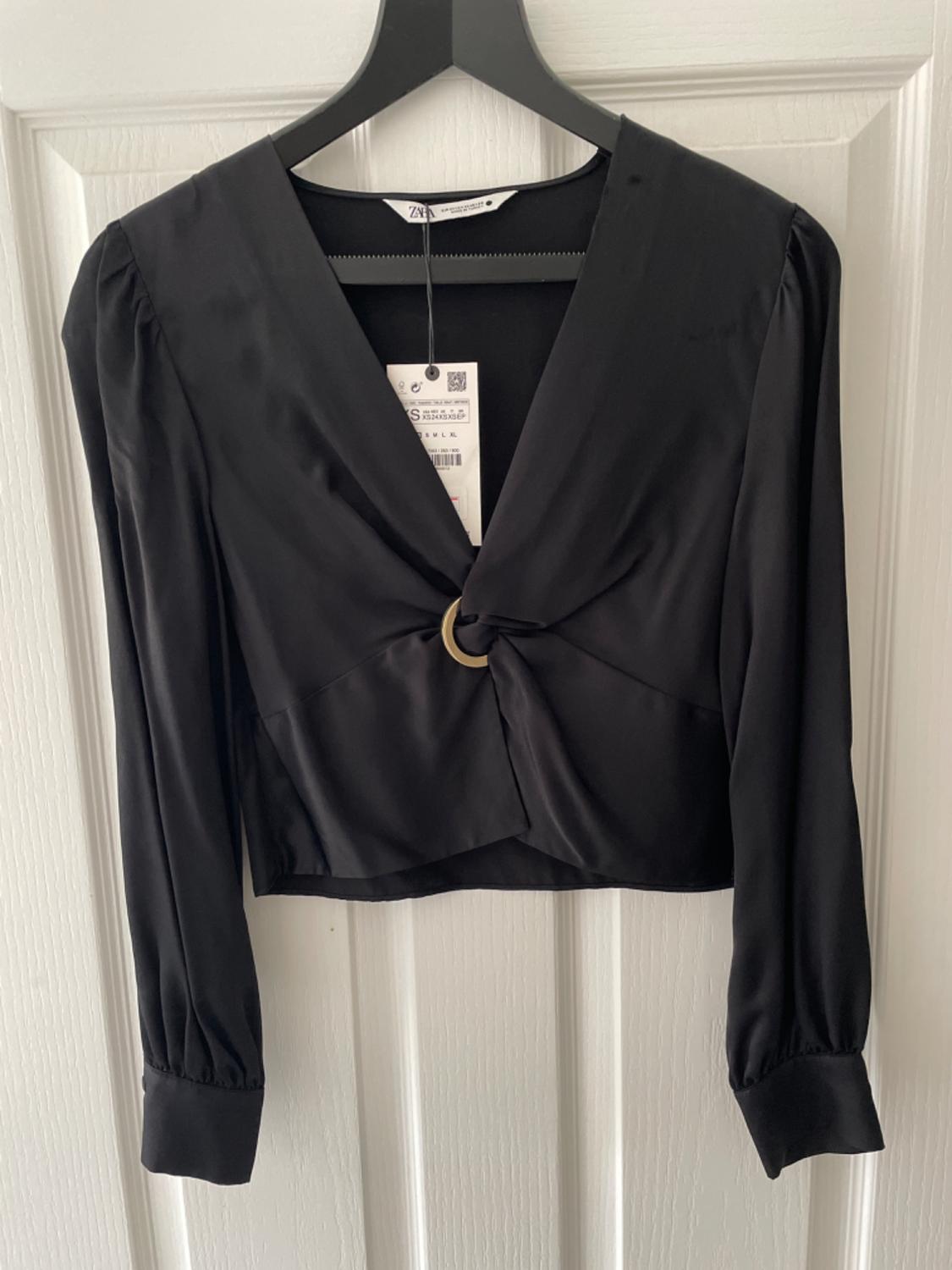 Black long-sleeved blouse with a V-neck and gold ring detail, hanging on a coat hanger over a white door. Label with the text ZARA.