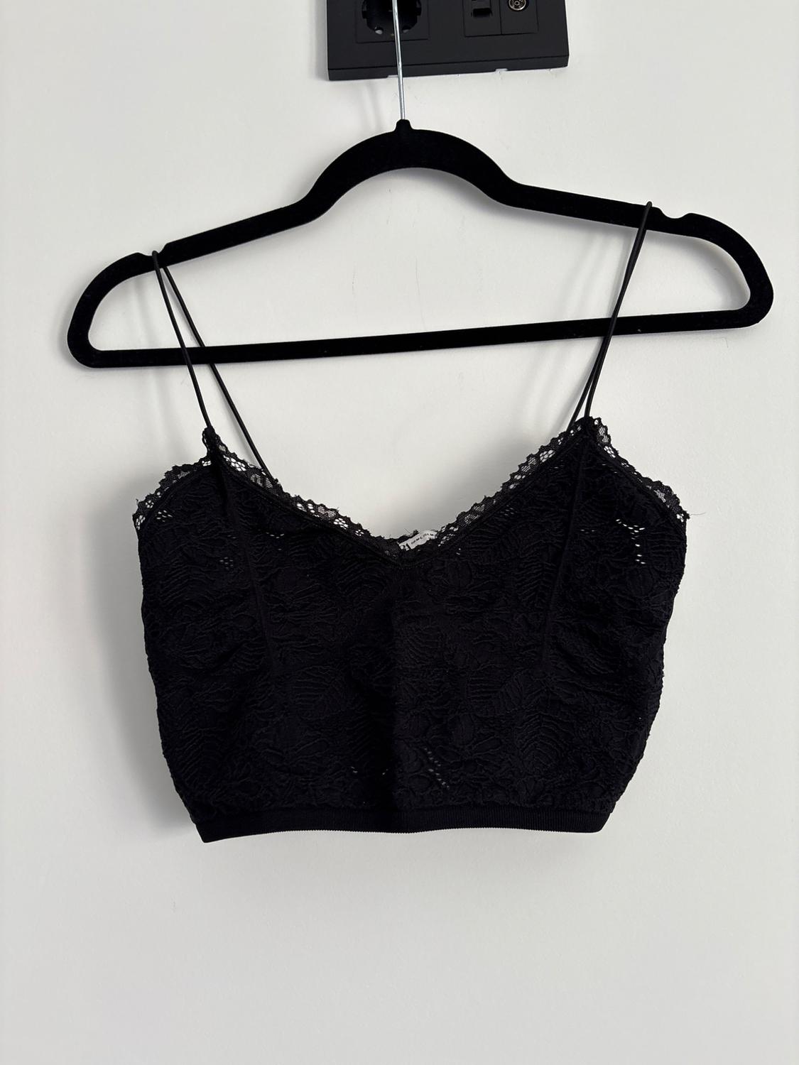 Black lace crop top with spaghetti straps and a ribbed hem, hanging on a black hanger against a white wall. Minimalist style.