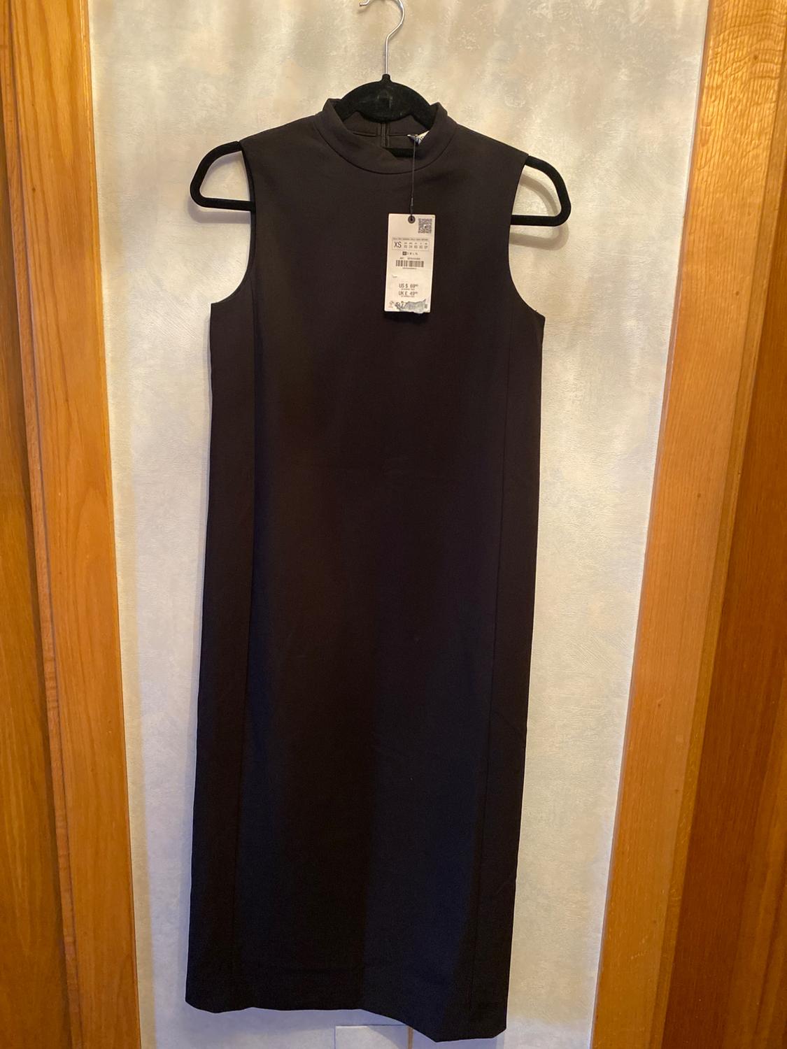 Sleeveless black dress hanging on a hanger, with a visible label in the centre. Light wall background and wooden frame.
