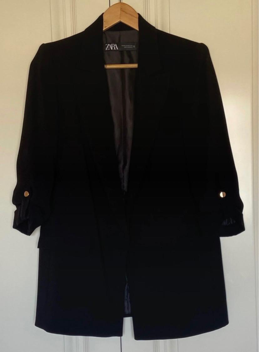Black jacket hanging on a wooden hanger, with three-quarter sleeves and gold buttons. Visible label with the text "ZARA".