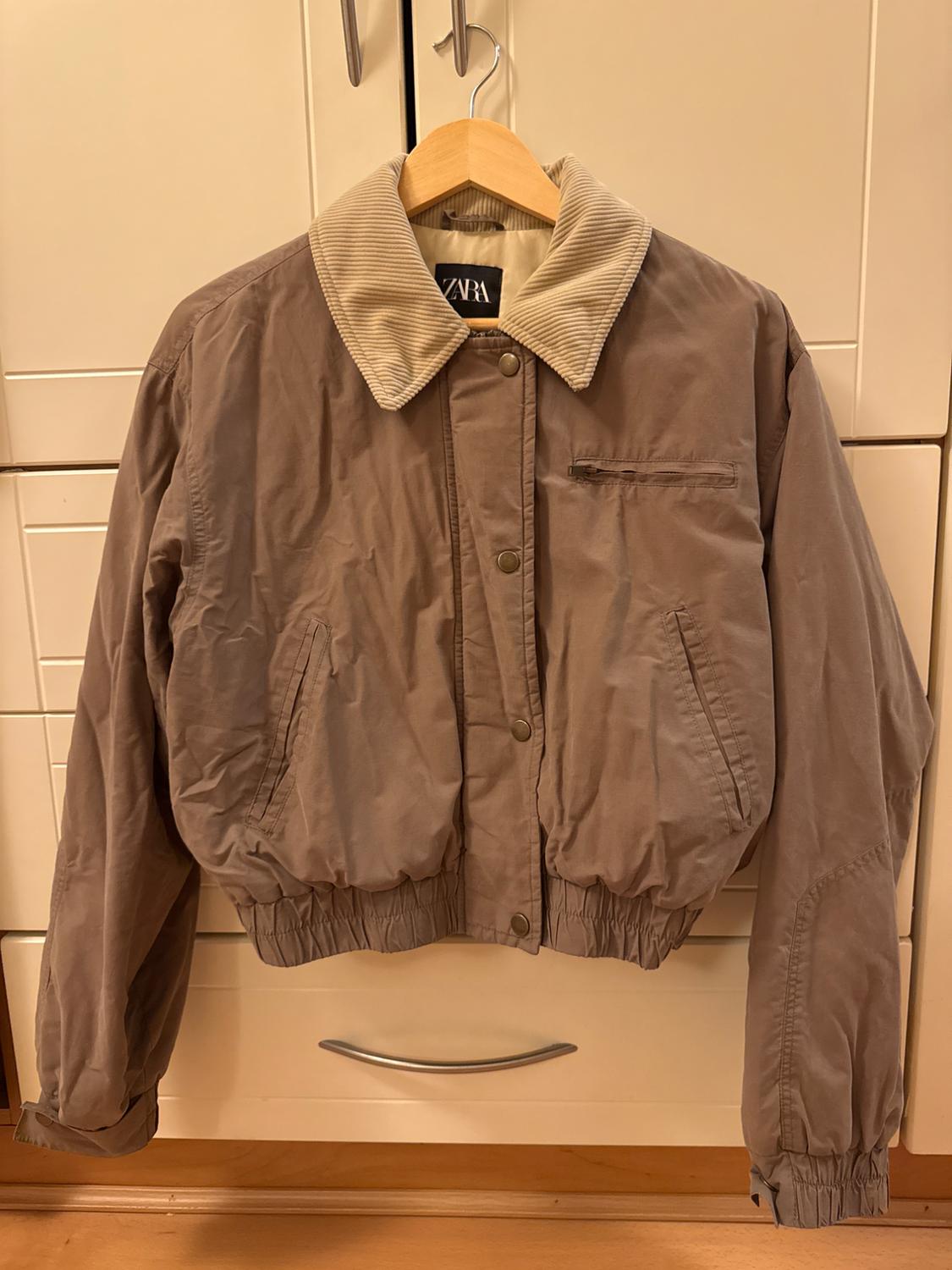 Light brown jacket with beige corduroy collar hanging on a wooden coat hanger. Visible label with the text "ZARA". Drawers in the background.