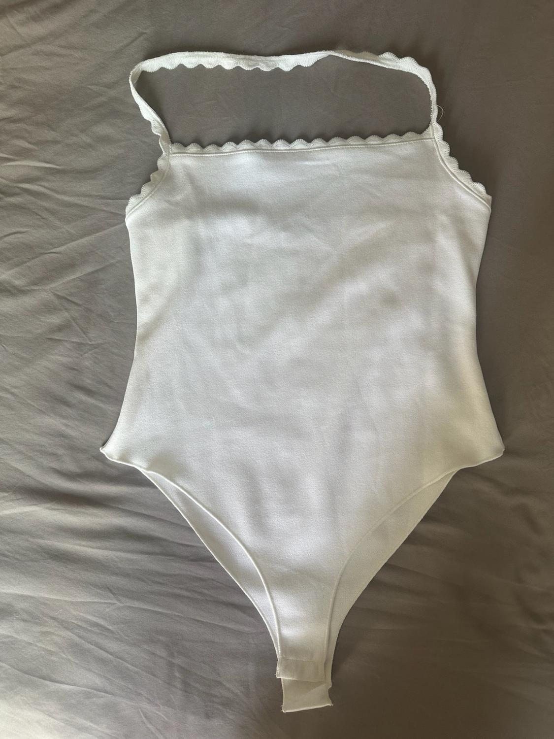 White halter-neck bodysuit with a scalloped trim, laid out on a crumpled grey sheet.