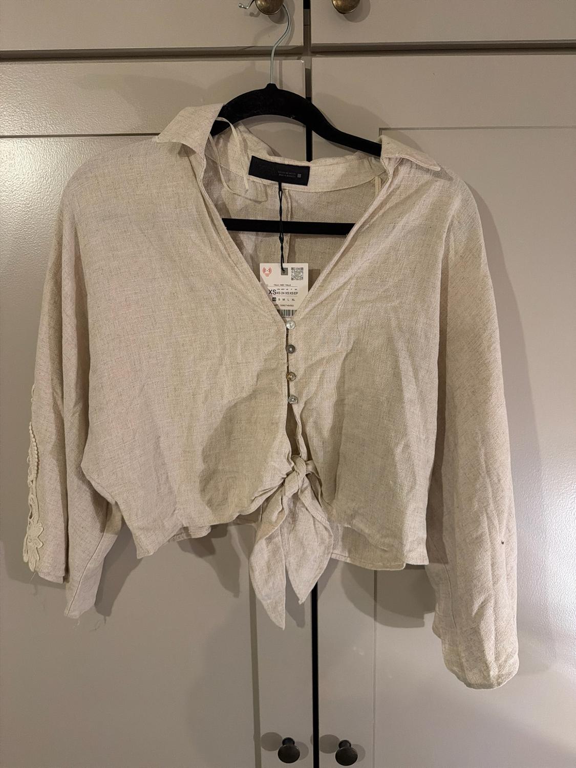 Beige shirt with buttons, a knot at the hem, lace detail and labels, hanging on a black hanger in front of wardrobe doors.