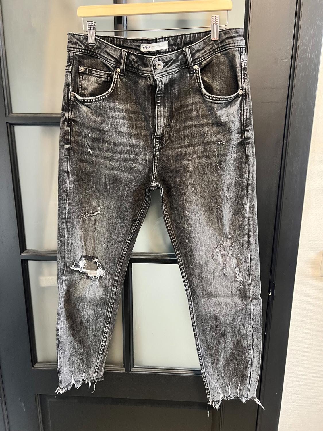 Worn grey jeans with rips at the knees, hanging on a wooden hanger in front of a black glass and wood door.