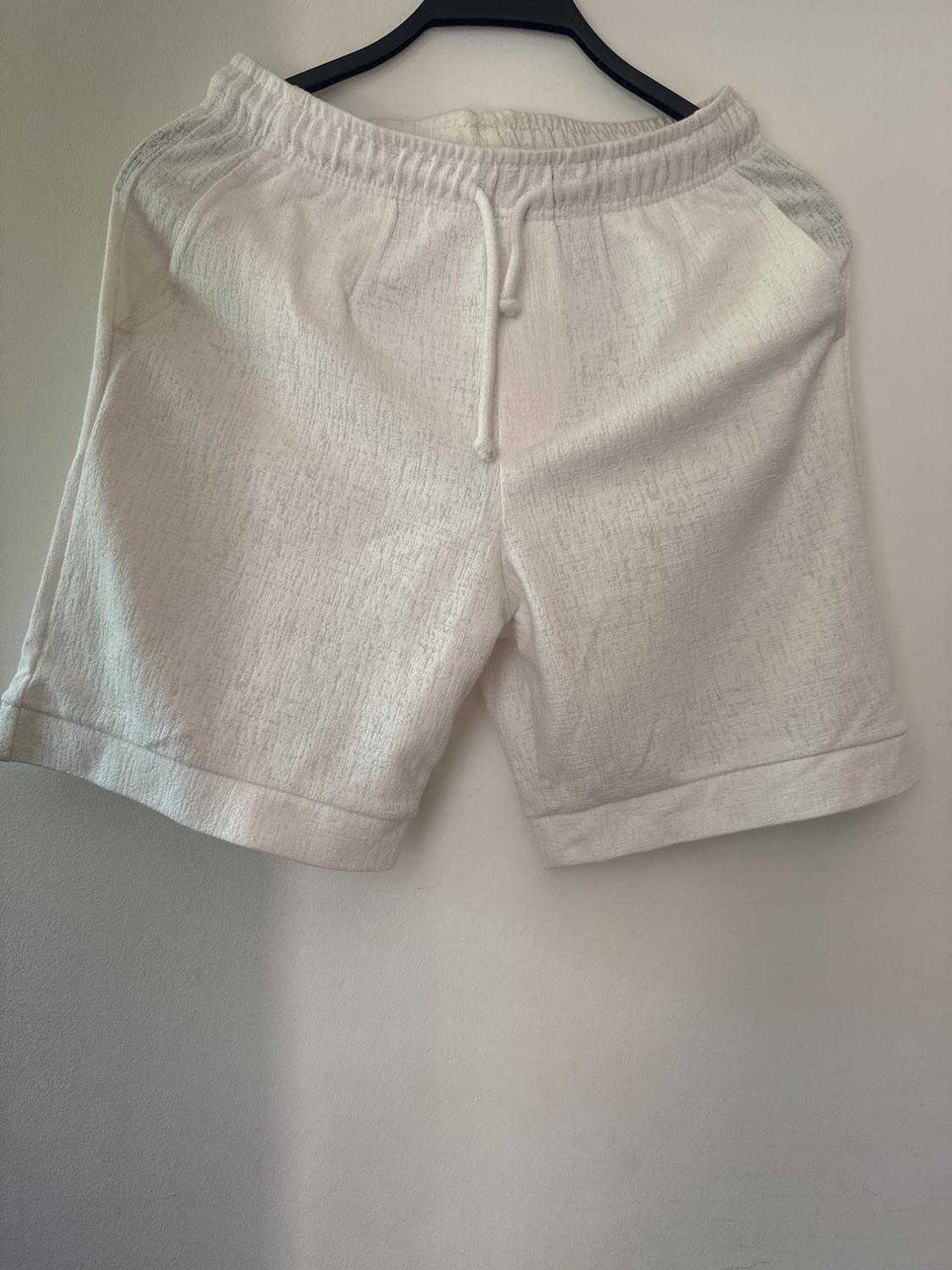 White, textured, relaxed-fit shorts with an elasticated drawstring waist, hanging on a black hanger against a white wall.