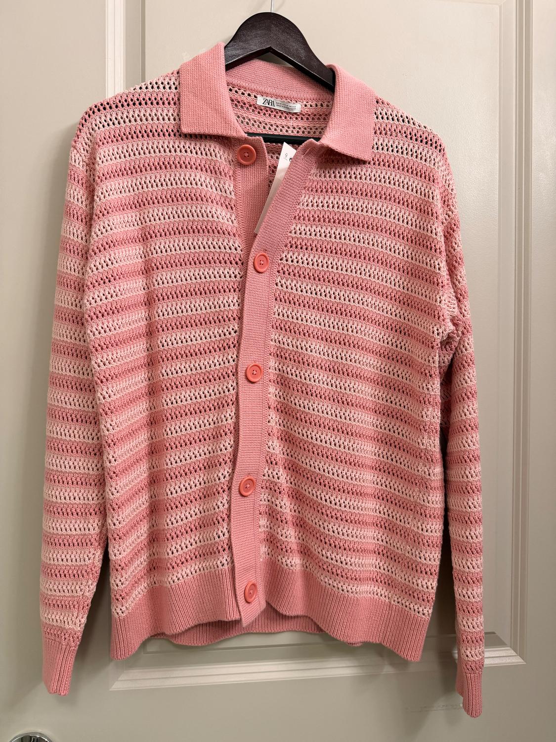 Pink knitted cardigan with stripes and large buttons, hanging on a black hanger over a beige door. Visible label on the collar.