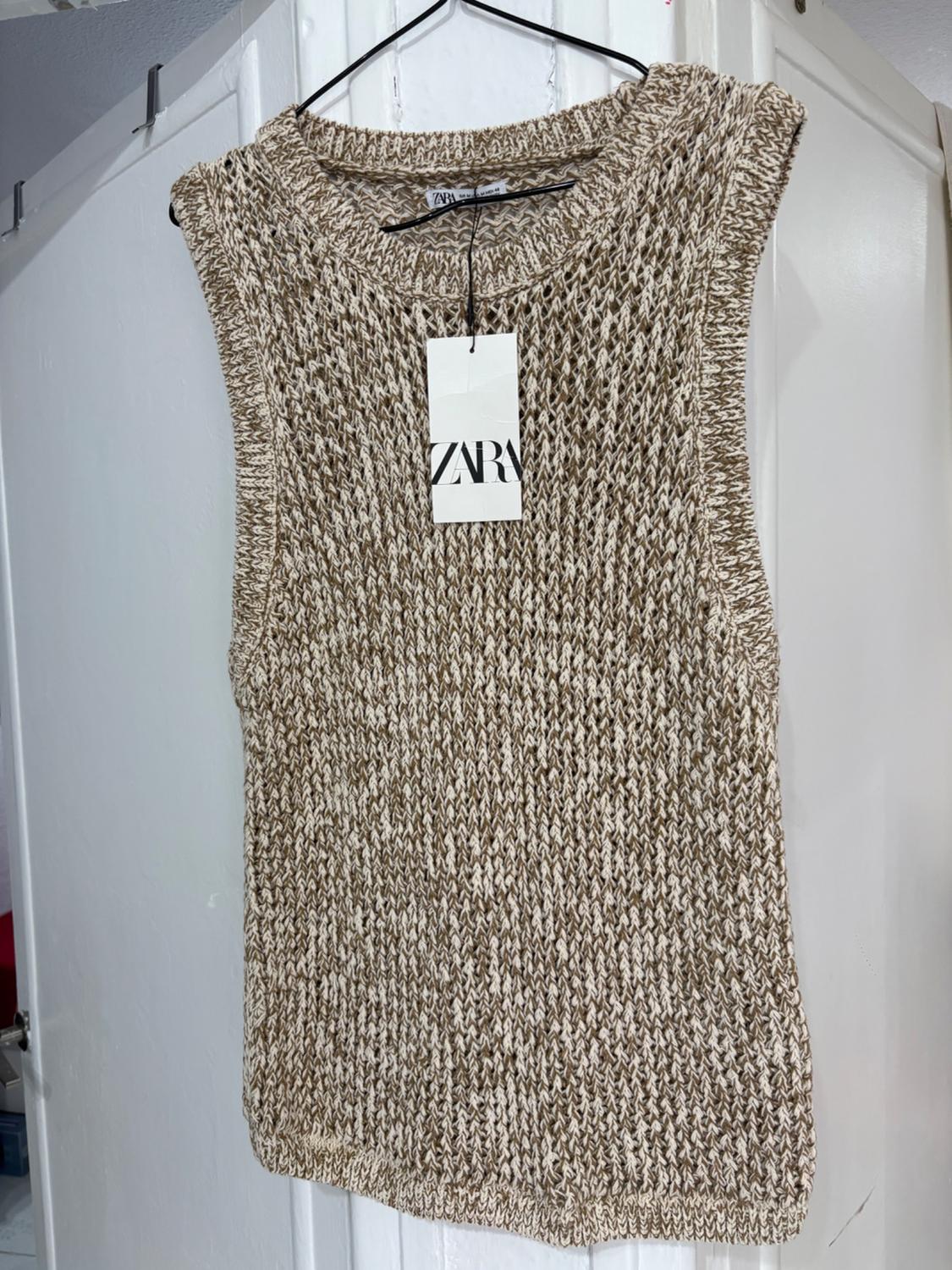 Knitted vest in beige and brown tones hanging on a hanger with a visible ZARA label, on a white background.