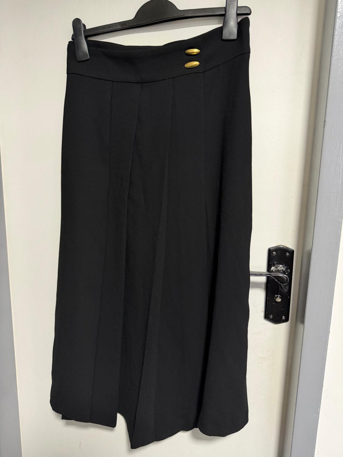 Long black skirt with two gold buttons at the waist, hanging on a black hanger over a white door.