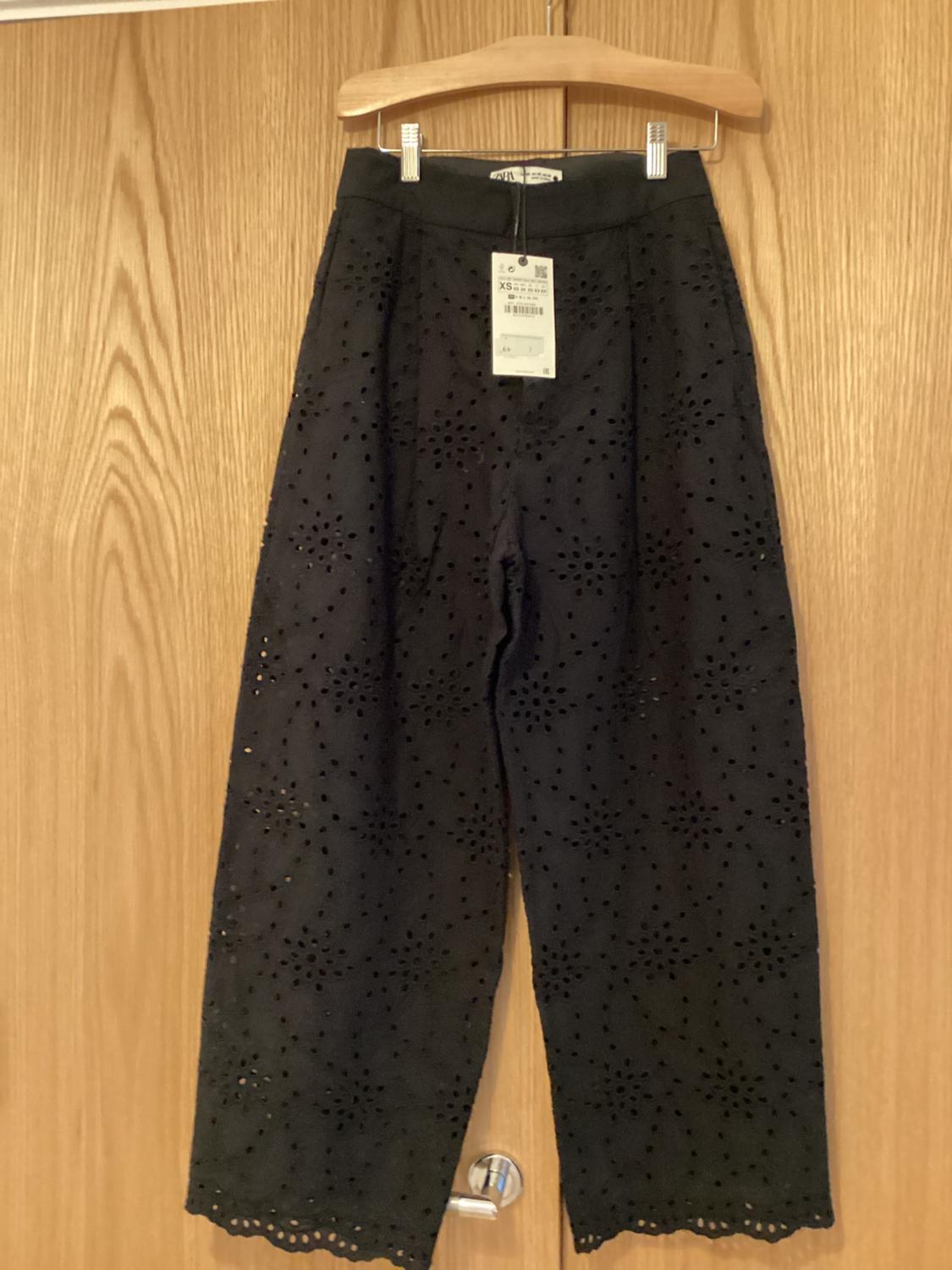 Black trousers with an openwork design hanging on a wooden hanger. Visible tag with product details. Wooden door in the background.
