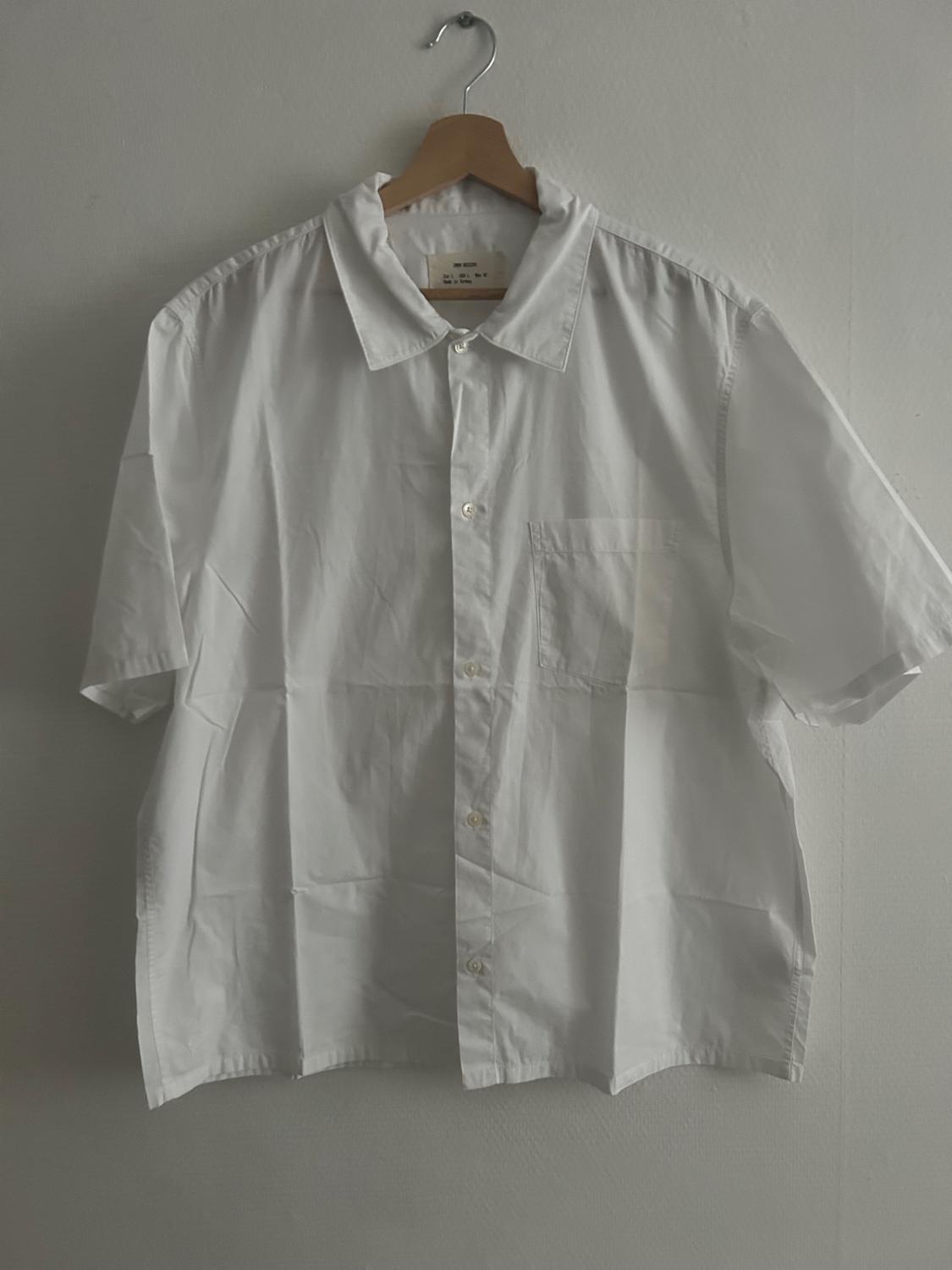 White short-sleeved shirt hanging on a wooden hanger against a grey wall. The label is visible inside the collar.