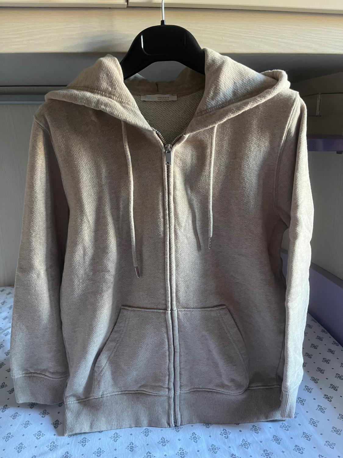 Casual beige hoodie with a zip and pockets, hanging on a black hanger over a bed with patterned sheets.