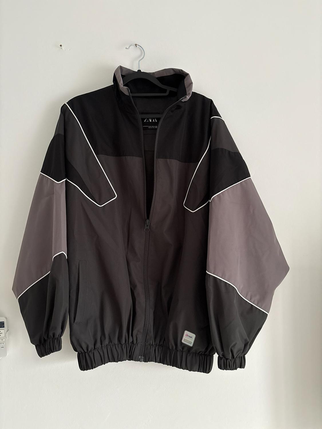 Black and grey colour-block jacket with white lines, hanging on a white wall. Text: ZARA, dnwr.