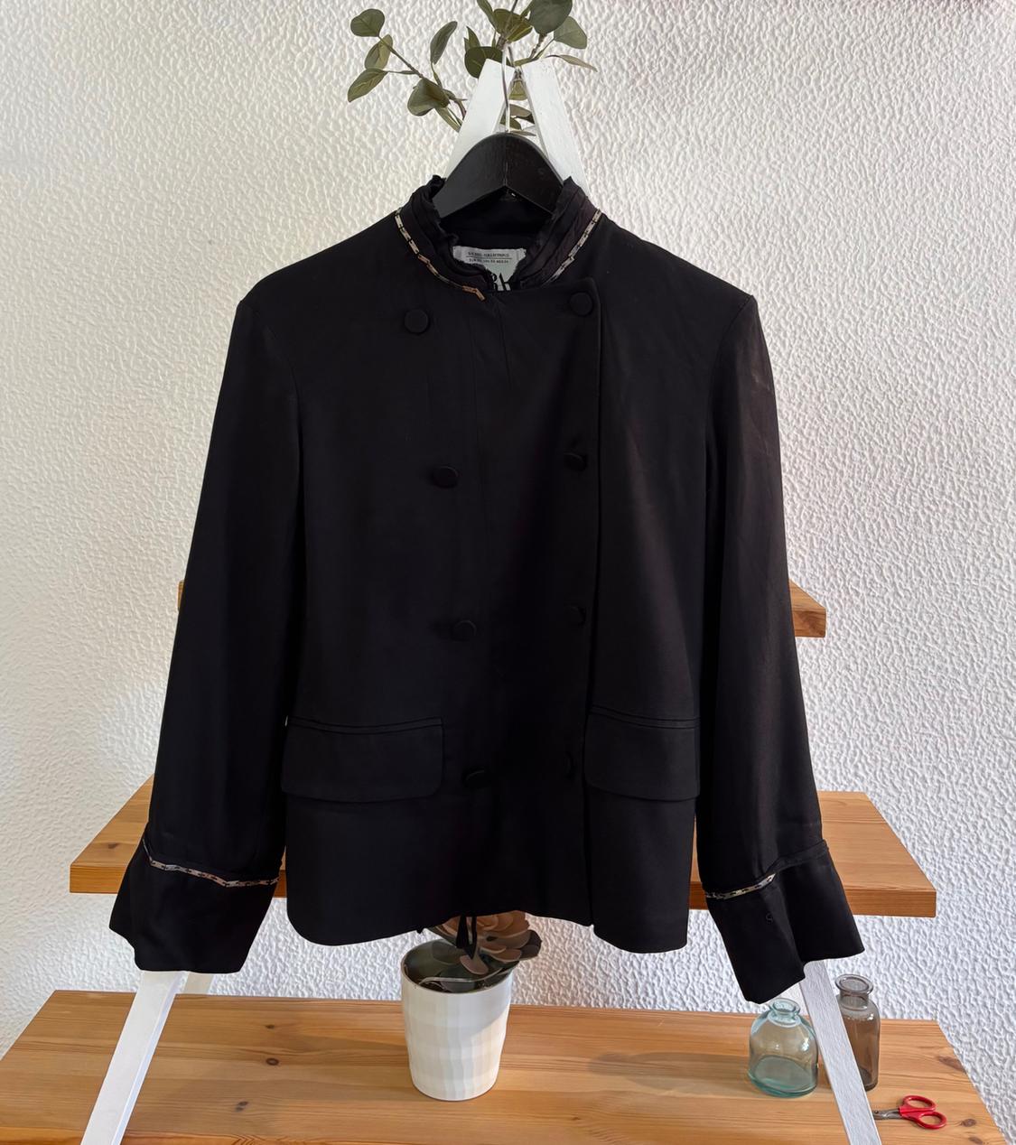 Black button-up jacket hanging on a white hanger against a textured wall background. There are plants and decorative jars.