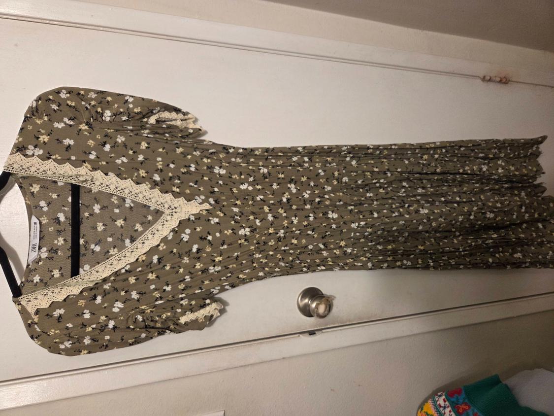 Long green floral print maxi dress, short sleeves and V-neck with cream lace, hanging on a black hanger on a white door.