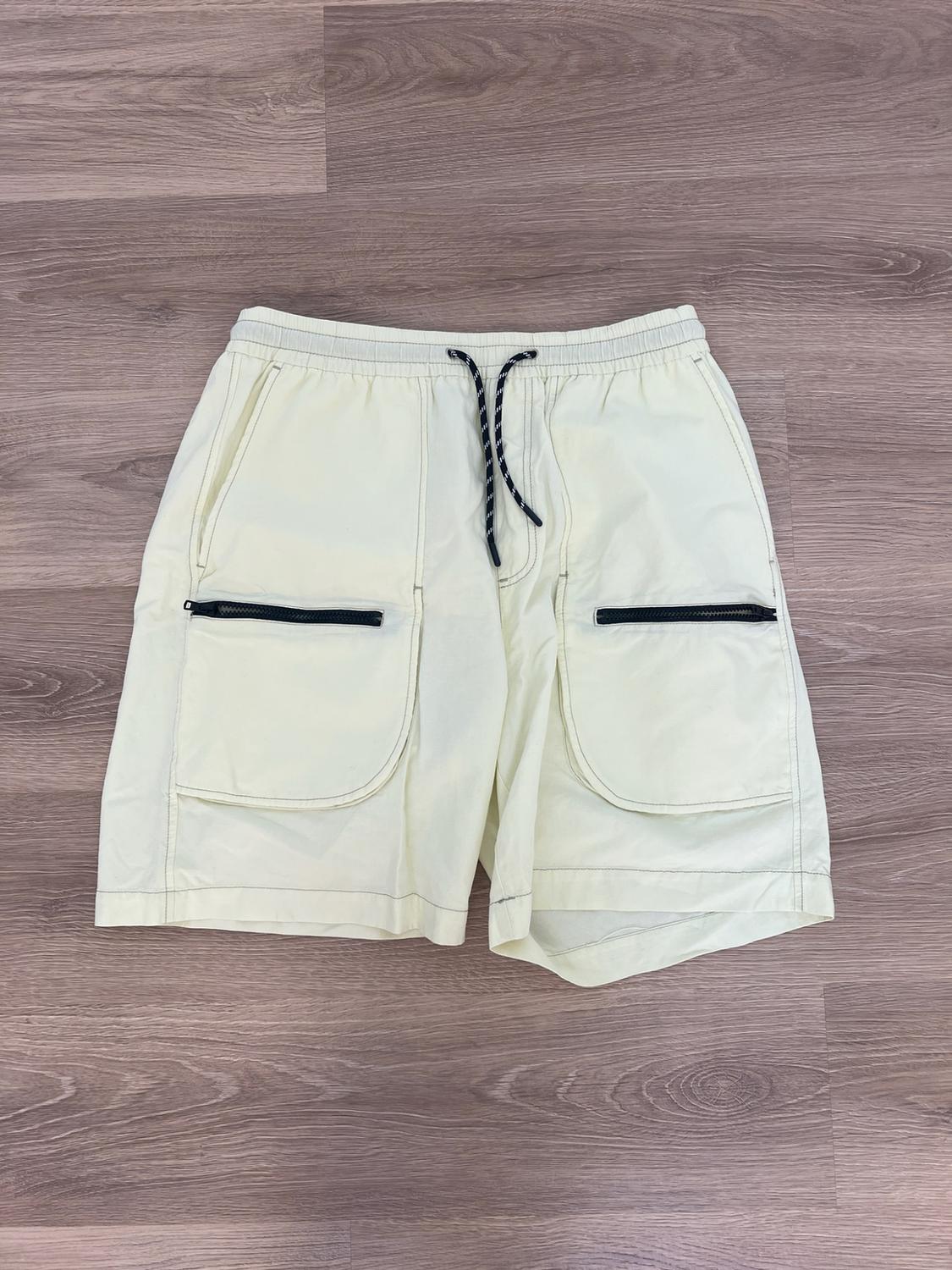Light yellow cargo-style shorts with an elasticated waist, drawstring and black zip pockets, on a wooden floor.
