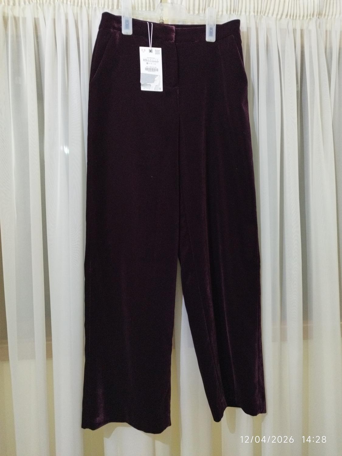 Dark-coloured velvet trousers hanging on a hanger in front of white curtains. Label visible. Date in image: 12/04/2026 14:28.