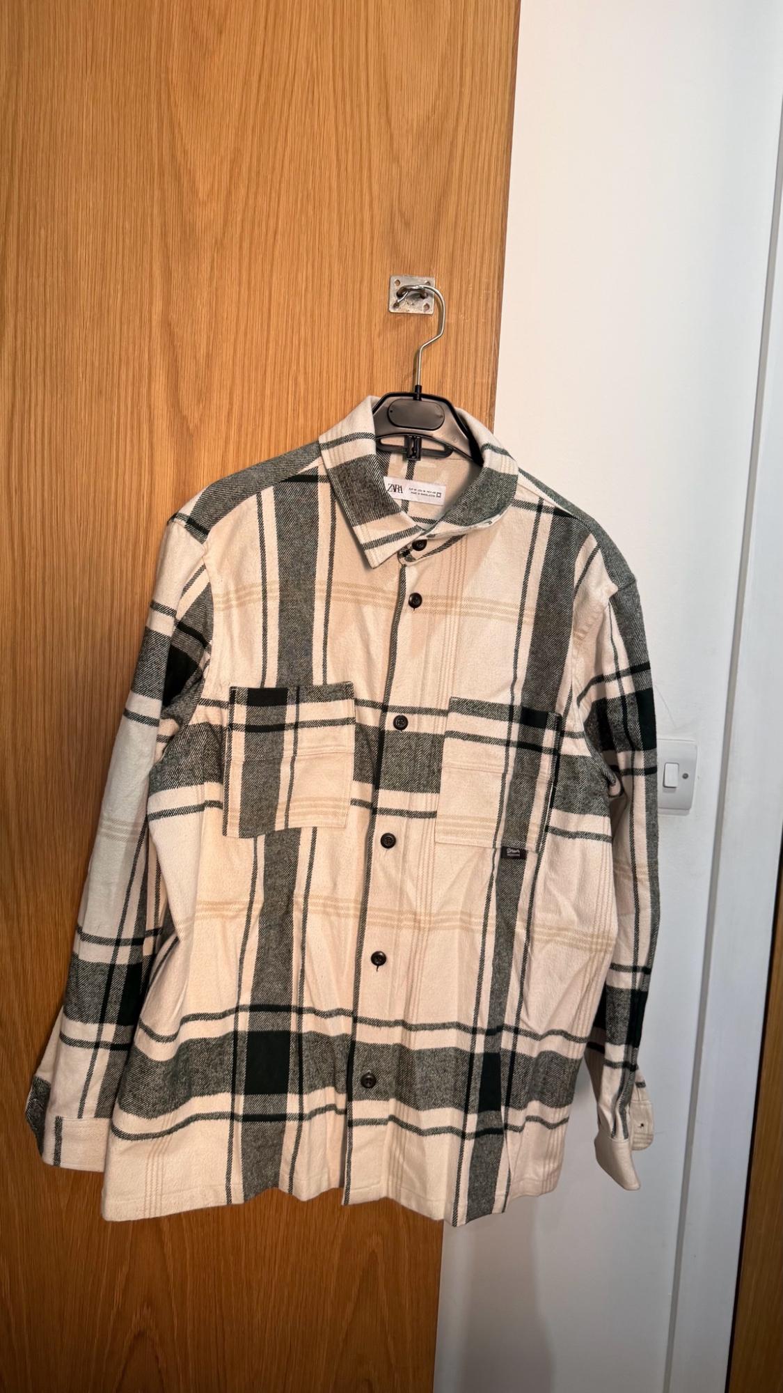 Long-sleeved checked shirt in cream and dark tones, hanging on a wooden door. Label with the text ZARA on the collar.